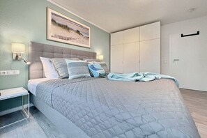A bedroom with a bed, bedside lamps, a painting, a wardrobe, and a door. - Holiday apartment Am Stadtwald 319611 (Kühlungsborn)