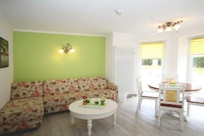 Una habitación con sofá modular, una mesita de centro con objetos decorativos, una mesa de comedor con sillas y una lámpara fijada a la pared.
