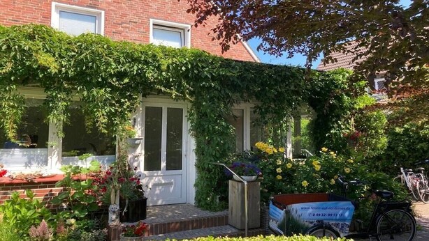 Een bakstenen gebouw met een witte deur, een kleine tuin met bloemen en planten, en een fiets voor de deur.
