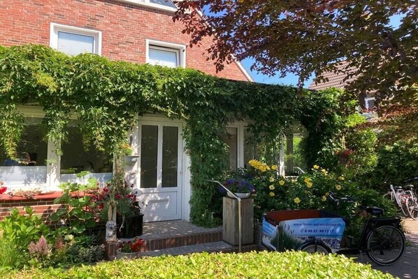 Un bâtiment en briques avec une porte blanche, un petit jardin avec des fleurs et des plantes, et un vélo garé à l’extérieur.