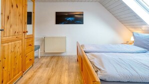 Een slaapkamer met een schuine kap, een houten garderobe, een bed met gestreept beddengoed, een nachtkastje, een aan de muur hangend schilderij en een raam.