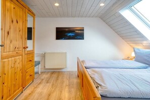 Une chambre avec un plafond en pente, une armoire en bois, un lit avec une literie à rayures, une table de chevet, un tableau accroché au mur et une fenêtre.