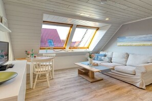 A loft-style room with a sloped ceiling, a dining area with a table and chairs, a sofa, and a window overlooking a red-roofed building. - Holiday apartment with 1 bedroom (Norderney)