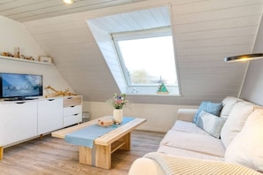 Ein moderner Loft-Wohnraum mit schräg versiegelter Decke, einem Fernseher auf einem weißen Schrank, einer Holzbank, einem beigefarbenen Sofa und einem Fenster mit Ausblick.