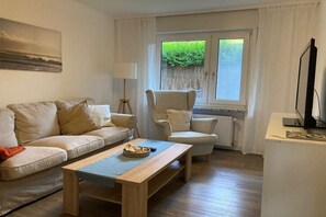 Ein Wohnzimmer mit Sofa, Sessel und Couchtisch, ein Fenster mit Blick auf Grünflächen und eine Heizung.
