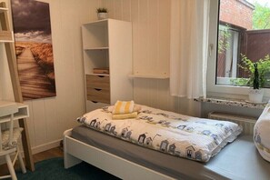 Une chambre avec un lit simple, une étagère, une fenêtre donnant sur un espace verdoyant et une étagère murale agrémentée d’une plante.