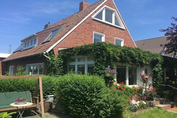 Une maison à deux étages, dotée d’un toit en pente, d’une façade en briques et d’un jardin où poussent diverses plantes et fleurs.