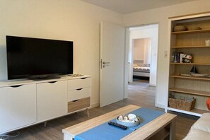 A modern room with a flat-screen TV on a white cabinet, a wooden coffee table with a remote control, and a bookshelf with various items. - Holiday apartment with 2 bedrooms (Norderney)