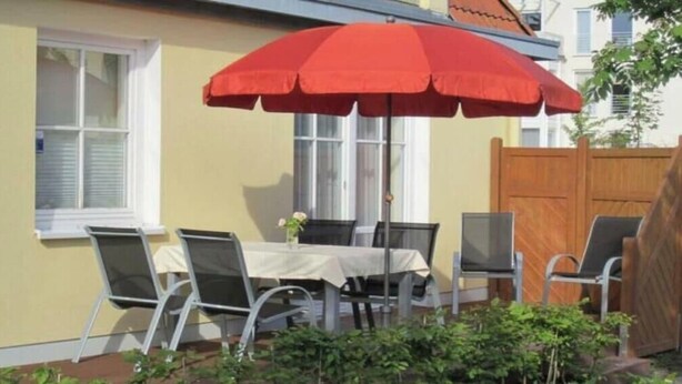 Uma área de refeições ao ar livre com mesa, cadeiras, um guarda-sol vermelho e um cercado de madeira.