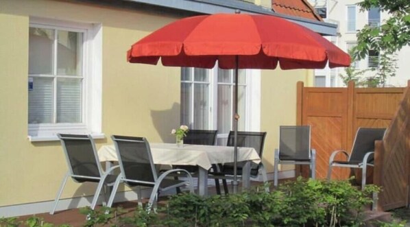 Un área de comedor al aire libre con mesa y sillas, una sombrilla roja y una valla de madera.
