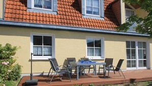 Uma casa com telhado de telhas, varanda com mesa e cadeiras e um jardim florido.