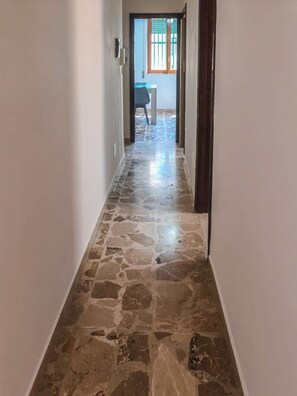 Un passadís amb sòl de pedra que condueix a una habitació amb finestra i una cadira.