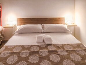 Cama bem arrumada com roupa de cama branca e um edredom marrom estampado, duas luminárias de cabeceira e uma cortina vermelha à esquerda.