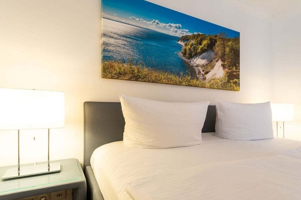 A hotel room with a bed, two bedside lamps, and a painting of a coastal landscape on the wall.