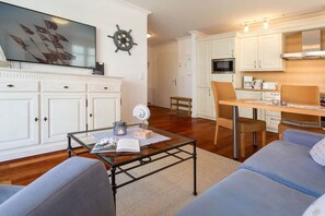 A living room with a television, a coffee table with a book and a candle, a dining area with a table and chairs, and a kitchen with cabinets and a microwave. - 3 (Beach Magic) in Villa Helene (Ostseebad Binz)