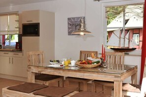 A dining area with a wooden table, chairs, and a glass-top table. There is a window with curtains and a view of a building outside. - Holiday home with 2 bedrooms (Born auf dem Darß)