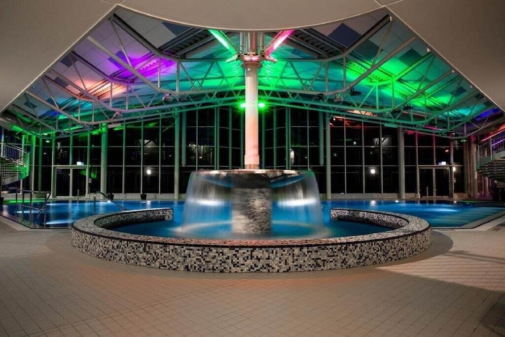 Piscina interior moderna com uma fonte central, rodeada por um espaço revestido em azulejos e um teto de vidro com vigas estruturais.