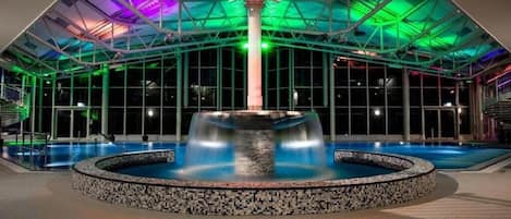A modern indoor pool with a central fountain, surrounded by a tiled area and a glass ceiling with structural beams.