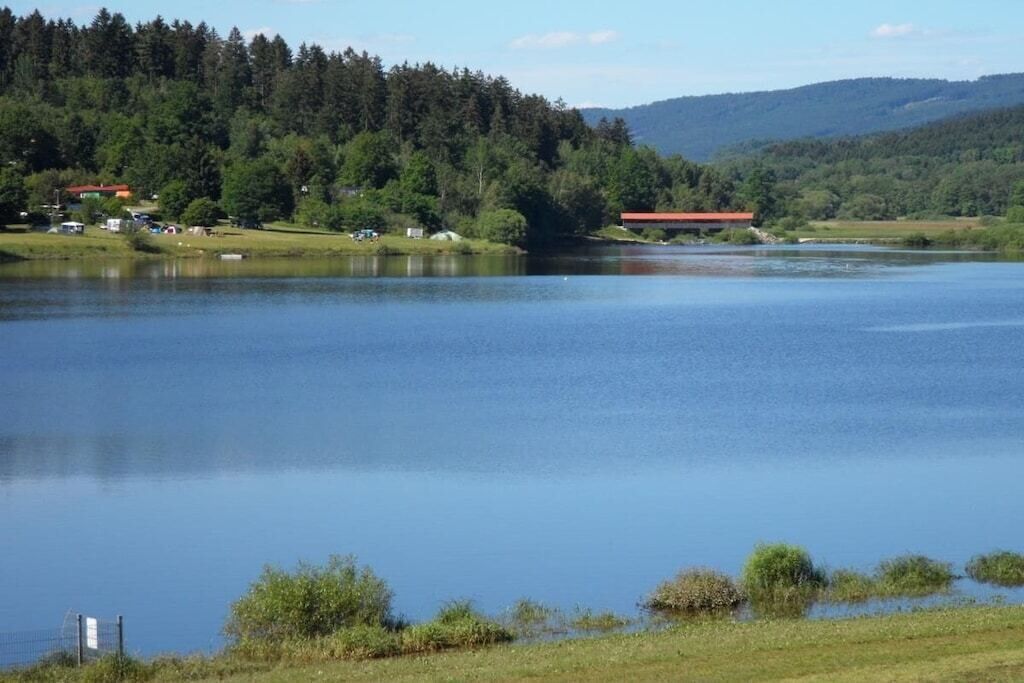 Un lago tranquilo con una colina boscosa al fondo y un edificio pequeño con techo rojo a la izquierda.