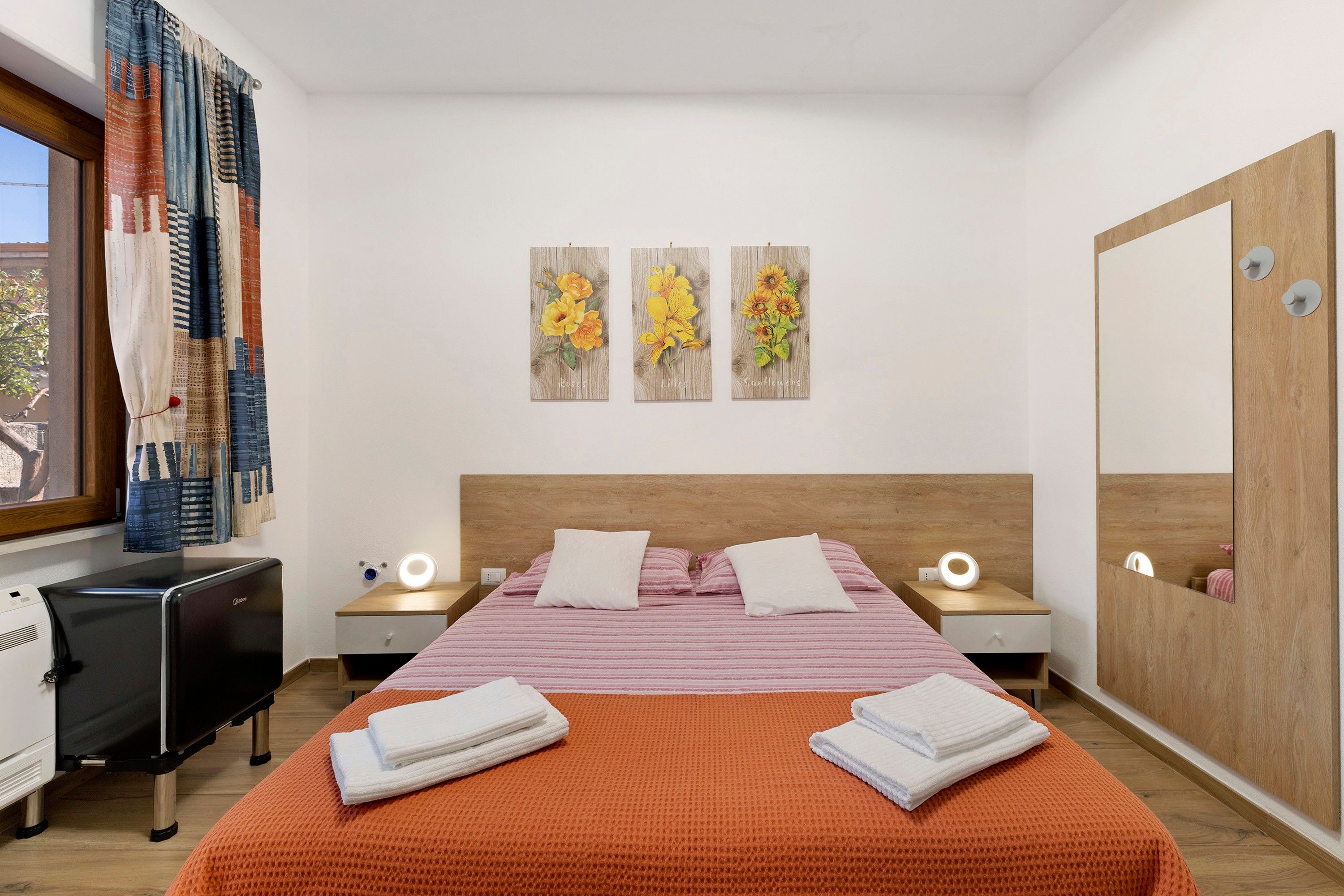 Ein Hotelzimmer mit einem Bett, zwei Nachttischen, einem Schreibtisch und einem Sessel. Über dem Bett hängen drei Wandbilder.