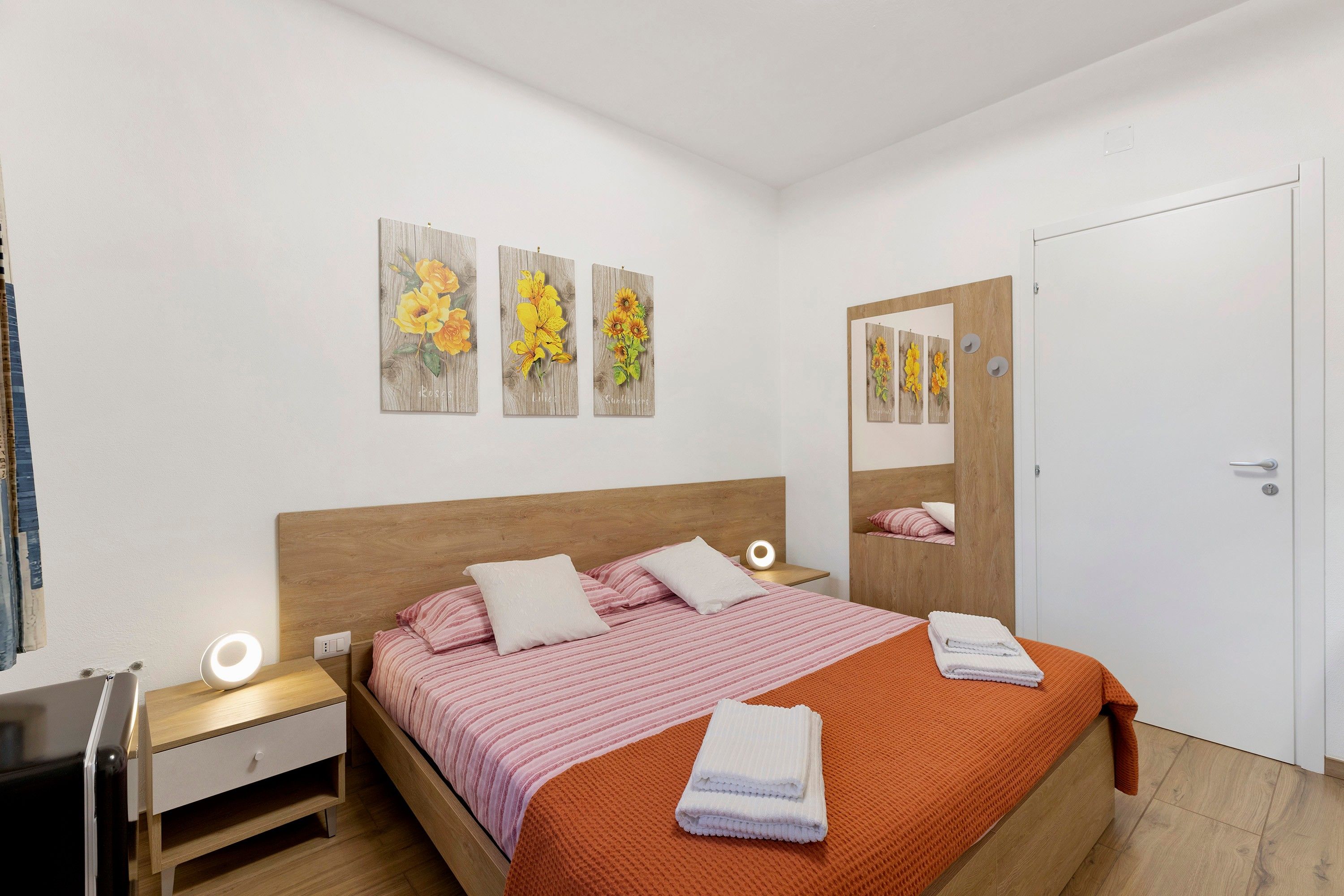 Ein Schlafzimmer mit einem hölzernen Kopfteil, einem Bett mit rosa-weiß gestreifter Bettwäsche, einem Nachttisch mit Lampe und einer Wand mit drei floralen Kunstwerken.