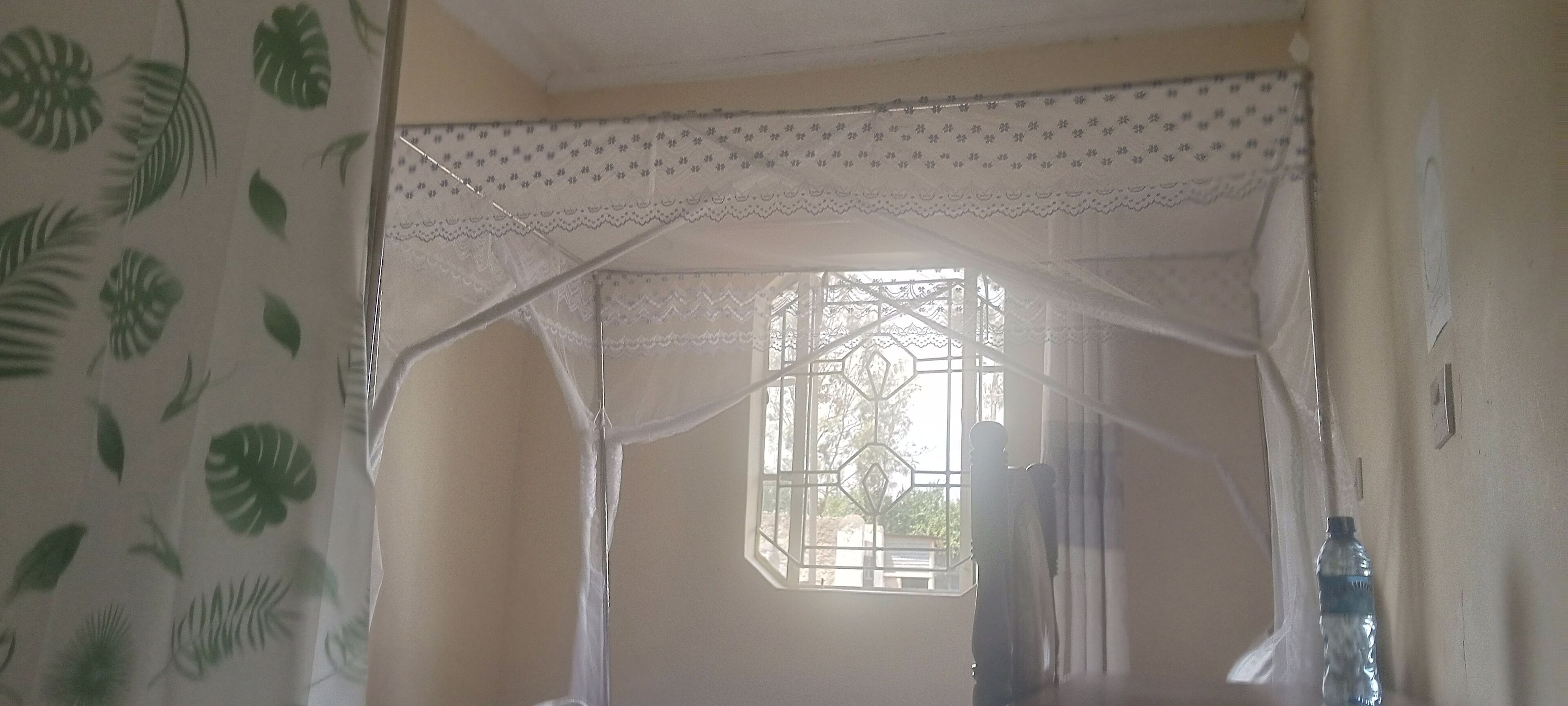 Quarto com cama de dossel, janela com padrão geométrico e uma garrafa de água ao lado.