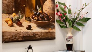 A painting of fruits and olives on a table, a vase with flowers, and a lantern on a table.