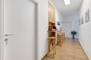 A hallway with a white door, a wooden cabinet, and a coffee machine. There are wooden stools and a vase with flowers.