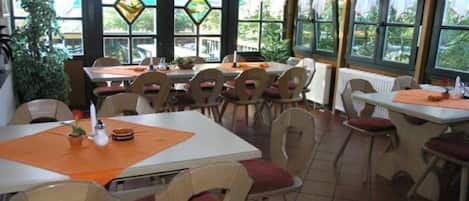 A dining area with multiple tables and chairs, large windows with stained glass, and a view of greenery outside.