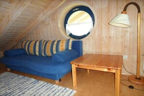 Ein Zimmer mit schrägem Dach, einem Rundfenster, einem blauen Sofa mit gestreiften Kissen, einem Holztisch und einer Stehlampe.
