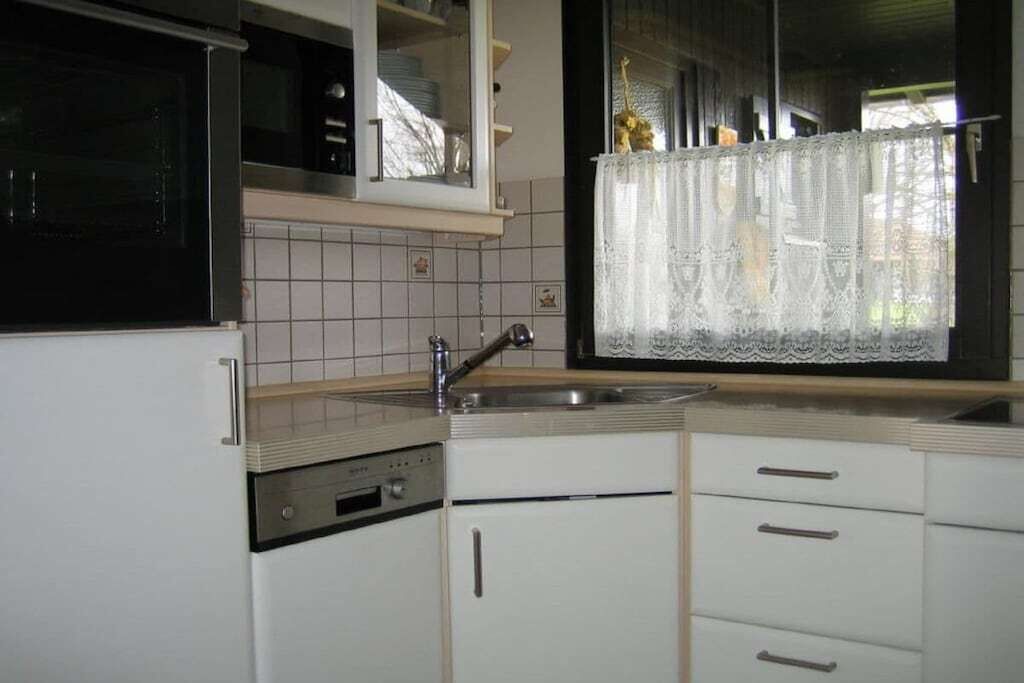 Eine Küche mit weißen Schränken, einer Edelstahlspüle und einem Fenster mit Spitze.