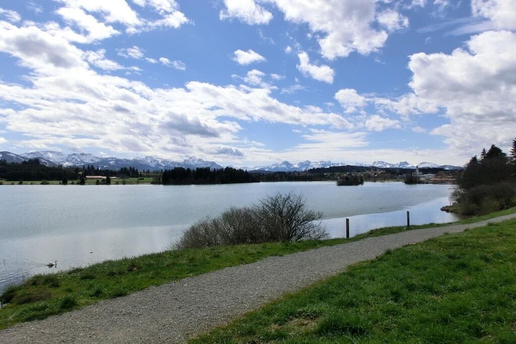 Um lago com uma trilha de cascalho e uma cerca, cercado por montanhas e árvores sob um céu parcialmente nublado.