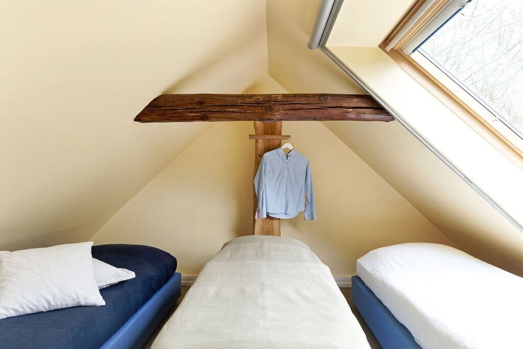 A small, triangular room with two beds, one with a blue mattress and the other with a white mattress. A wooden beam ceiling and a window with a view are visible.