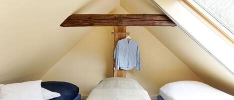 A small, triangular room with two beds, one with a blue mattress and the other with a white mattress. A wooden beam ceiling and a window with a view are visible.
