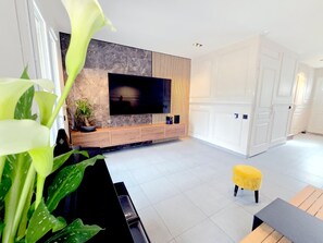 A modern living room with a flat-screen TV mounted on a marble wall, a wooden entertainment unit, and a yellow ottoman. - Holiday Home “Chez Elie!” with Private Terrace, Garden, and Wi-Fi (Orléans)