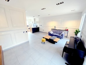 Una sala de estar moderna con un sofá, una mesa de centro y un pequeño otoman. Se observa una cocina al fondo con una lavadora.