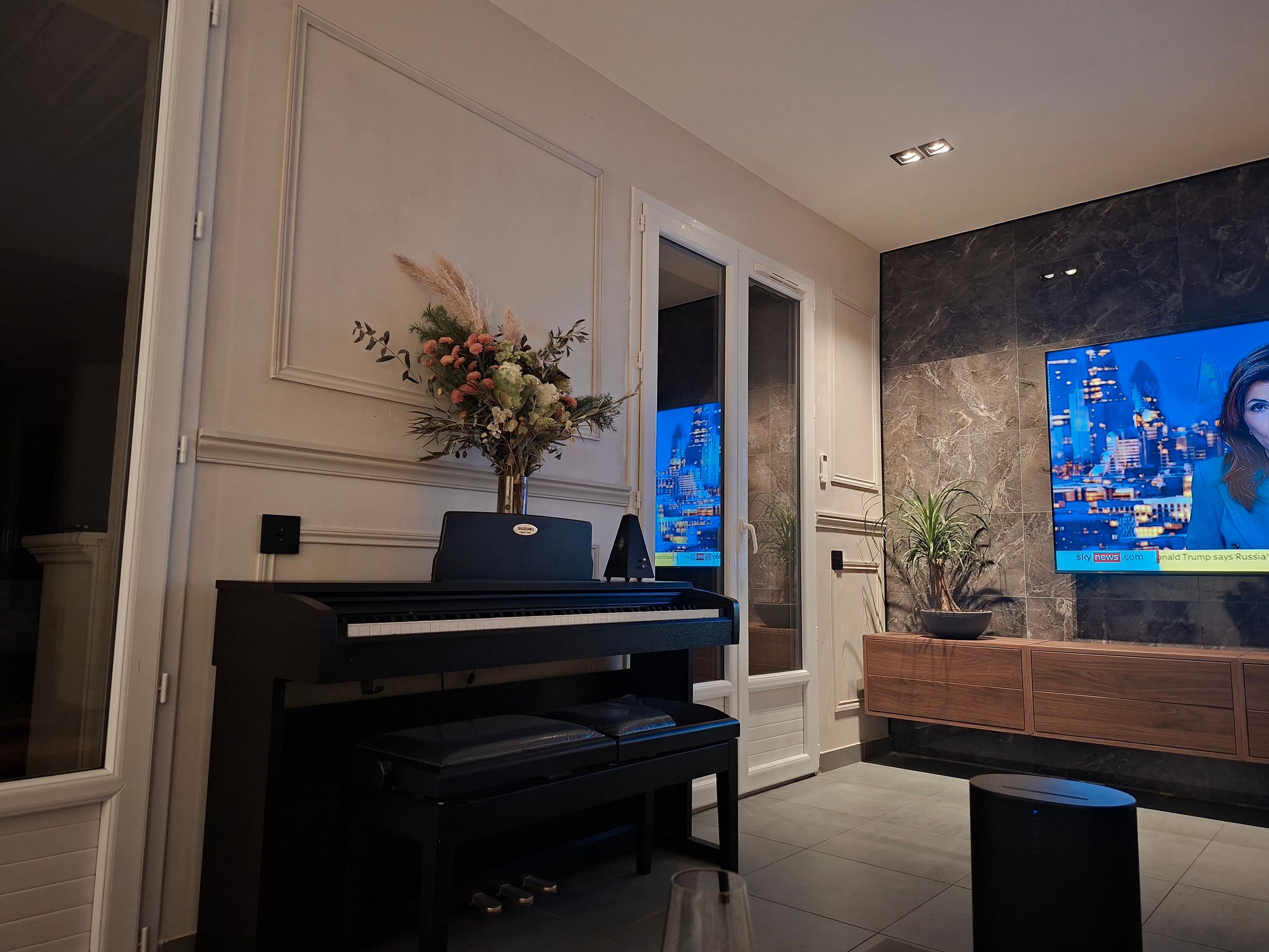 Een moderne woonkamer met een piano, een televisie waarop een nieuwskanaal wordt vertoond, en een decoratieve vaas met bloemen.