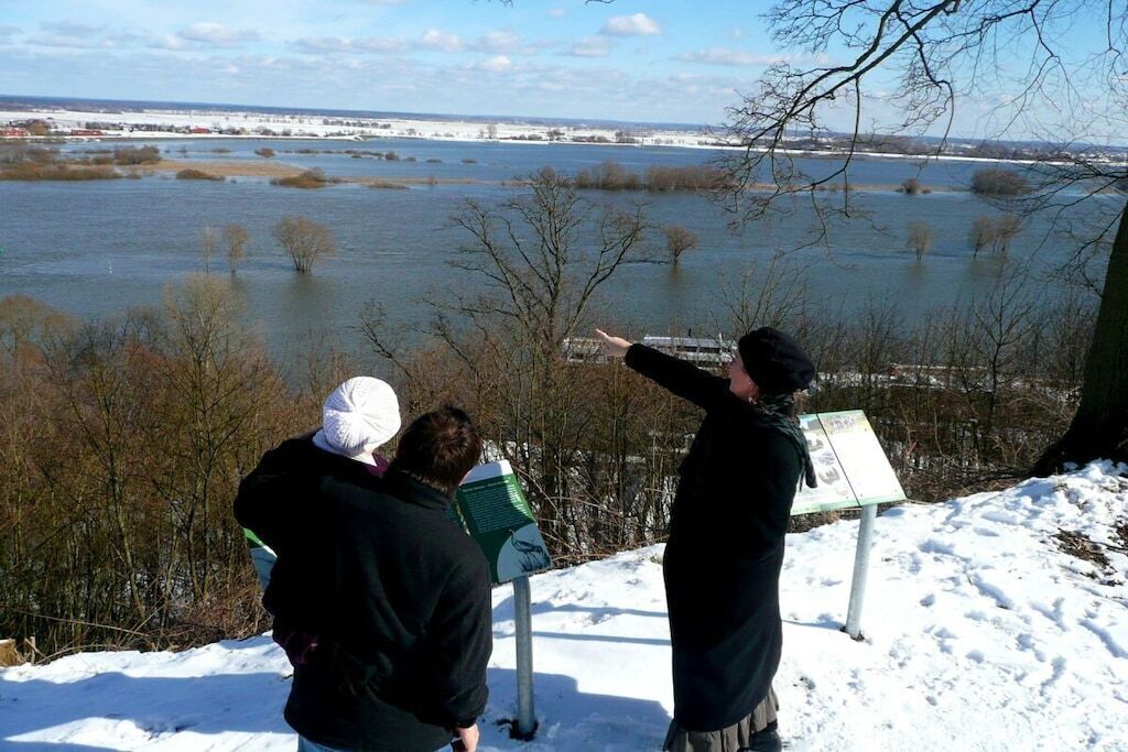 Un grupo de personas de pie junto a un cartel cerca de un cuerpo de agua, con nieve en el suelo.