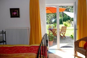 Una habitación con cama, una silla, una mesa y un ventanal corredero que da a un área exterior con patio y otra silla.