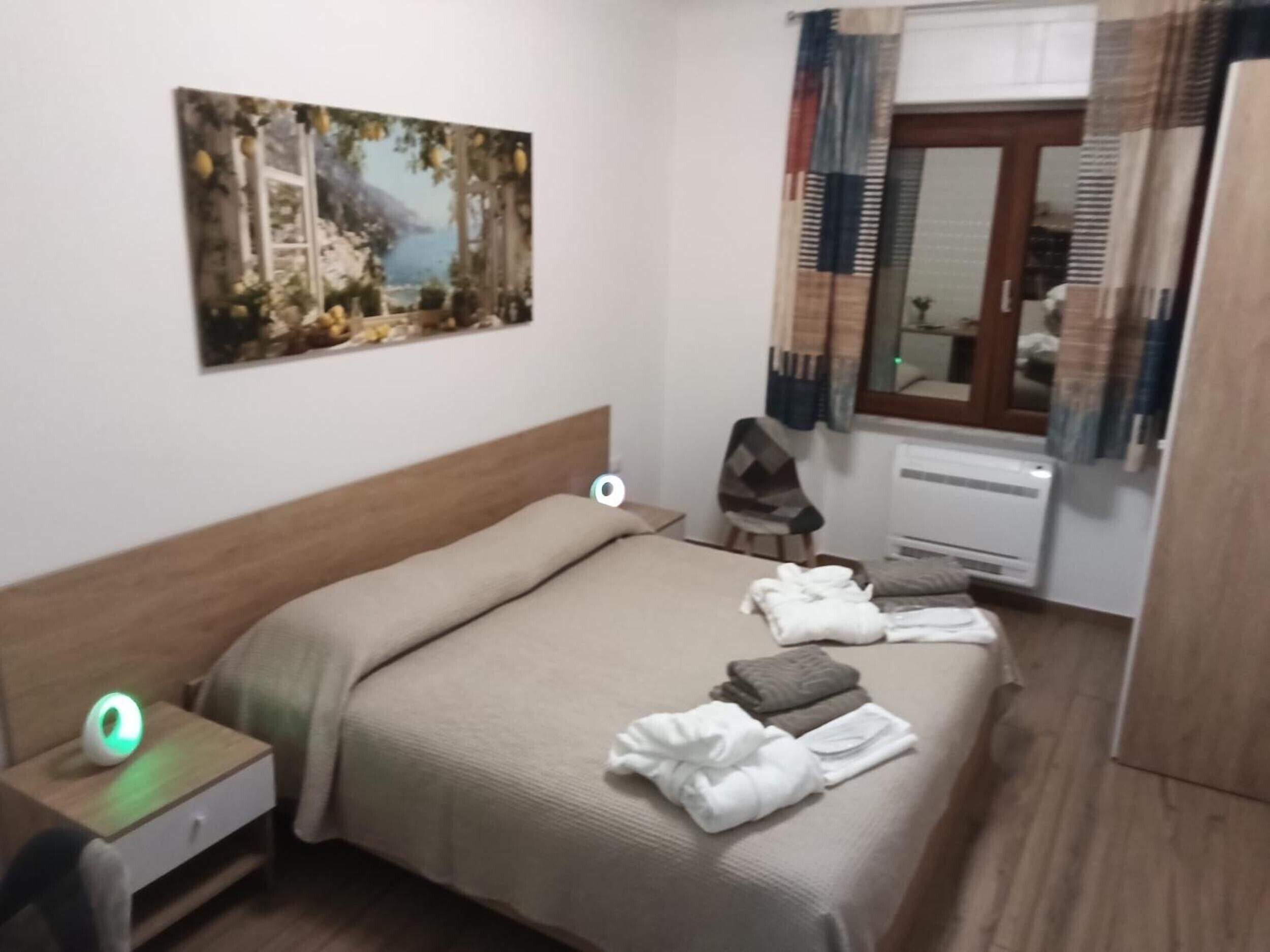 A hotel room with a bed, a bedside table with a lamp, a painting on the wall, and a window with curtains.