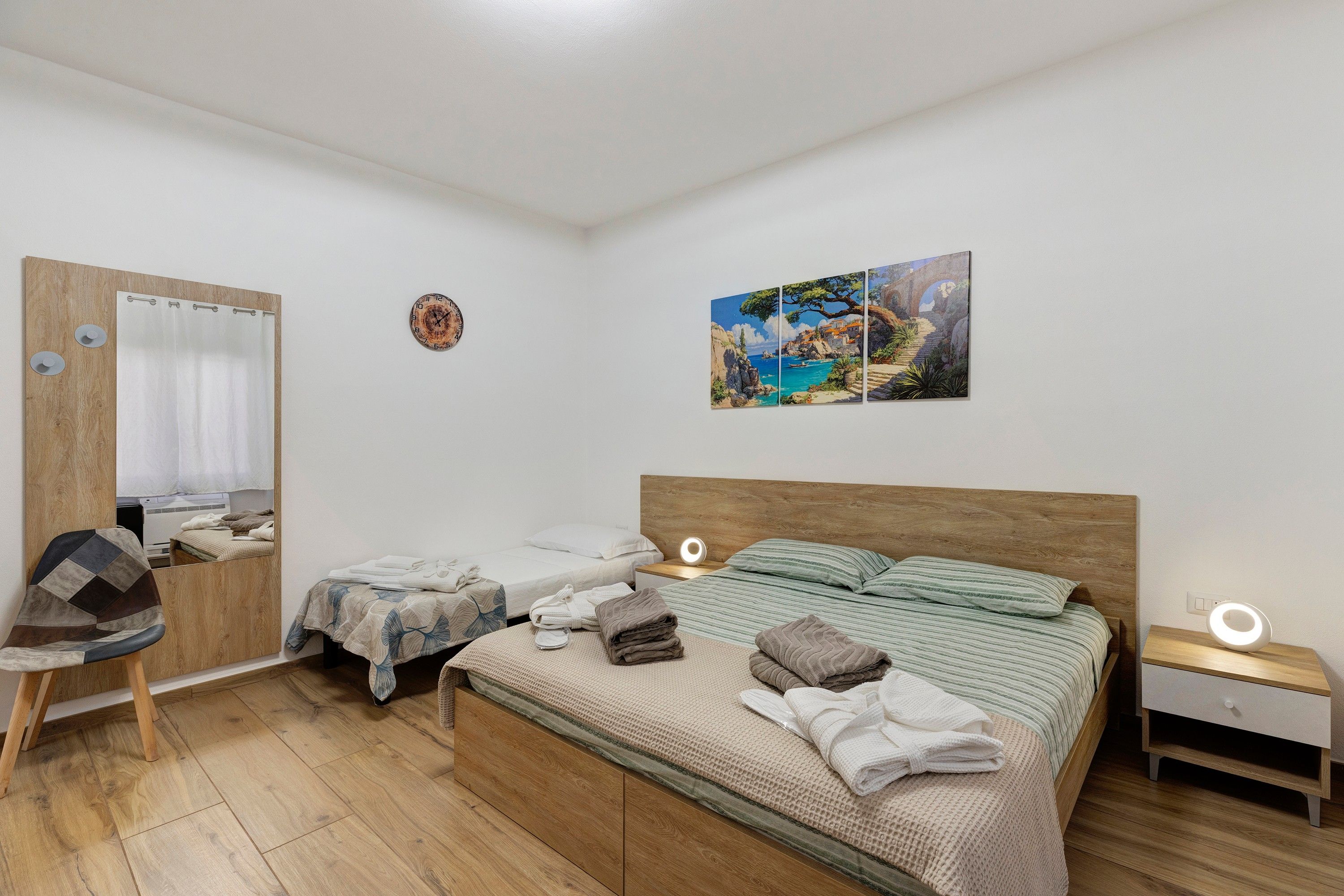 Un dormitorio con cabecera de madera, dos camas individuales, una silla de madera, una mesita de noche con una lámpara, un reloj de pared y un cuadro.