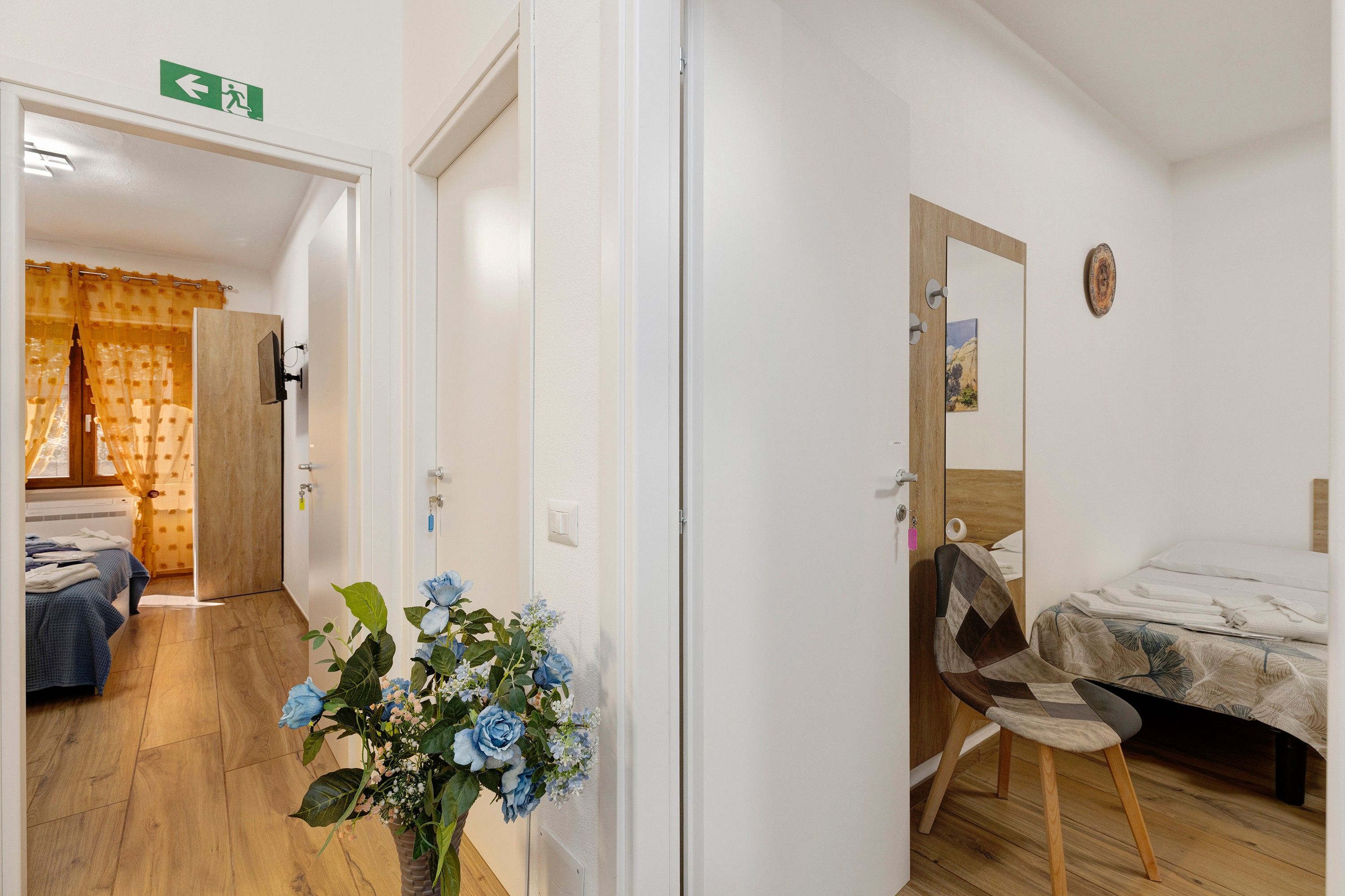 Une chambre d’hôtel avec un lit, une chaise, une petite table et un tableau accroché au mur. On y trouve une porte donnant sur une autre pièce et un interrupteur visible.