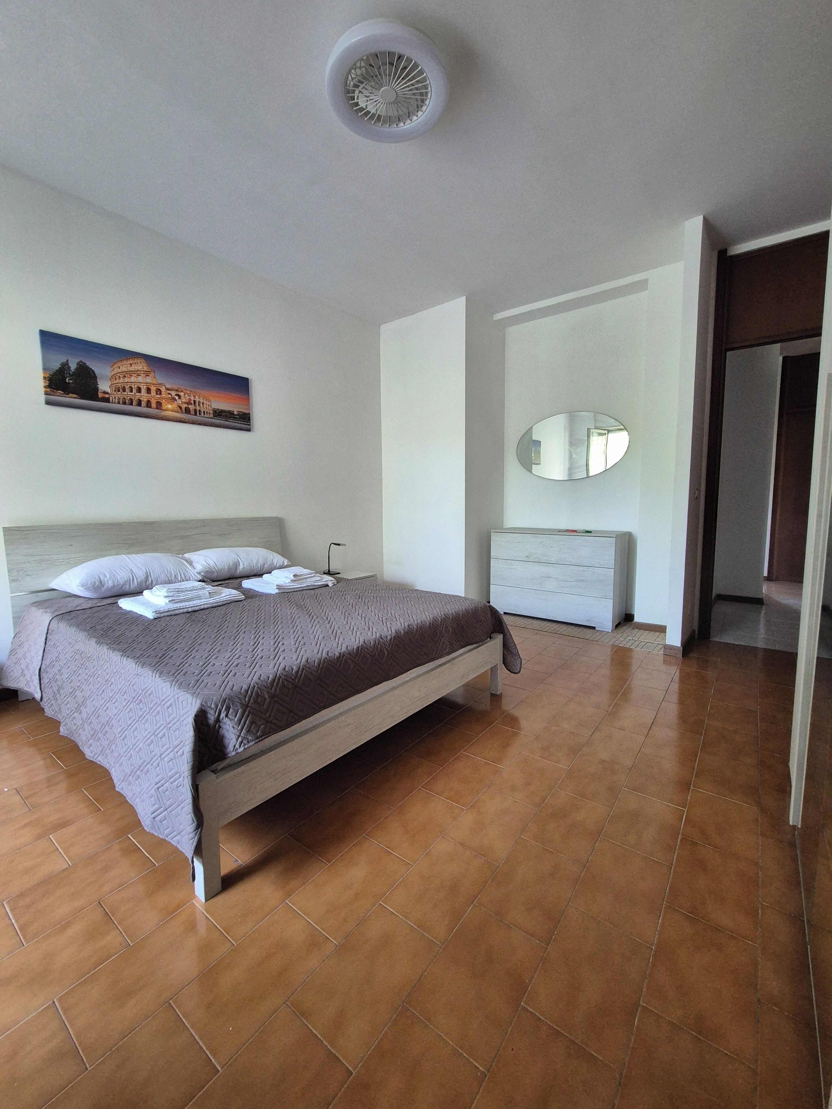 Une chambre à coucher comprenant un lit, une table de chevet, une commode, un miroir et un tableau représentant le Colisée.