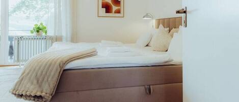 Een slaapkamer met een bed, een radiator, een raam met gordijnen en een wandlamp.