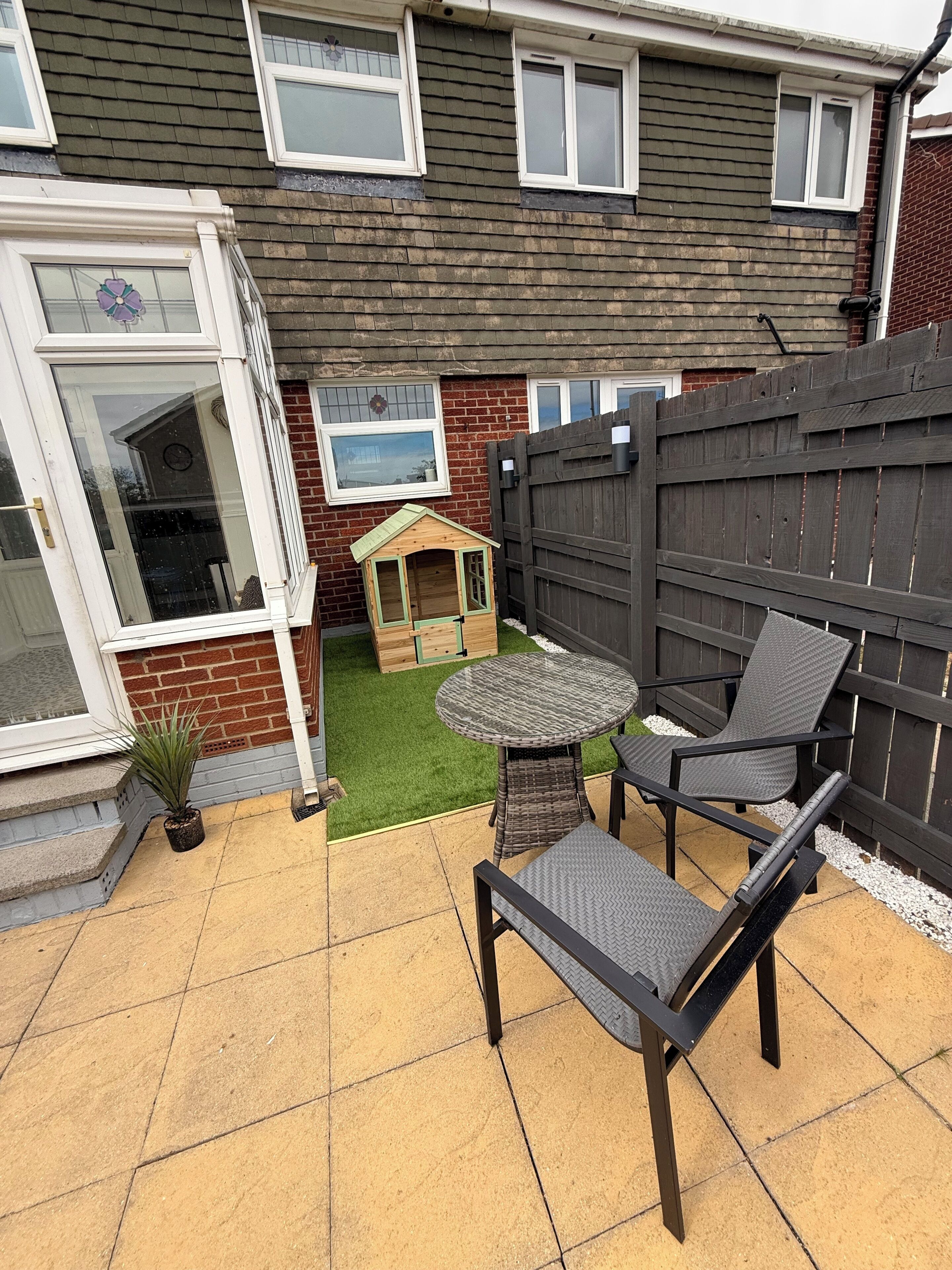 Une terrasse avec une table et des chaises, une petite maison de jeu et une clôture en bois.