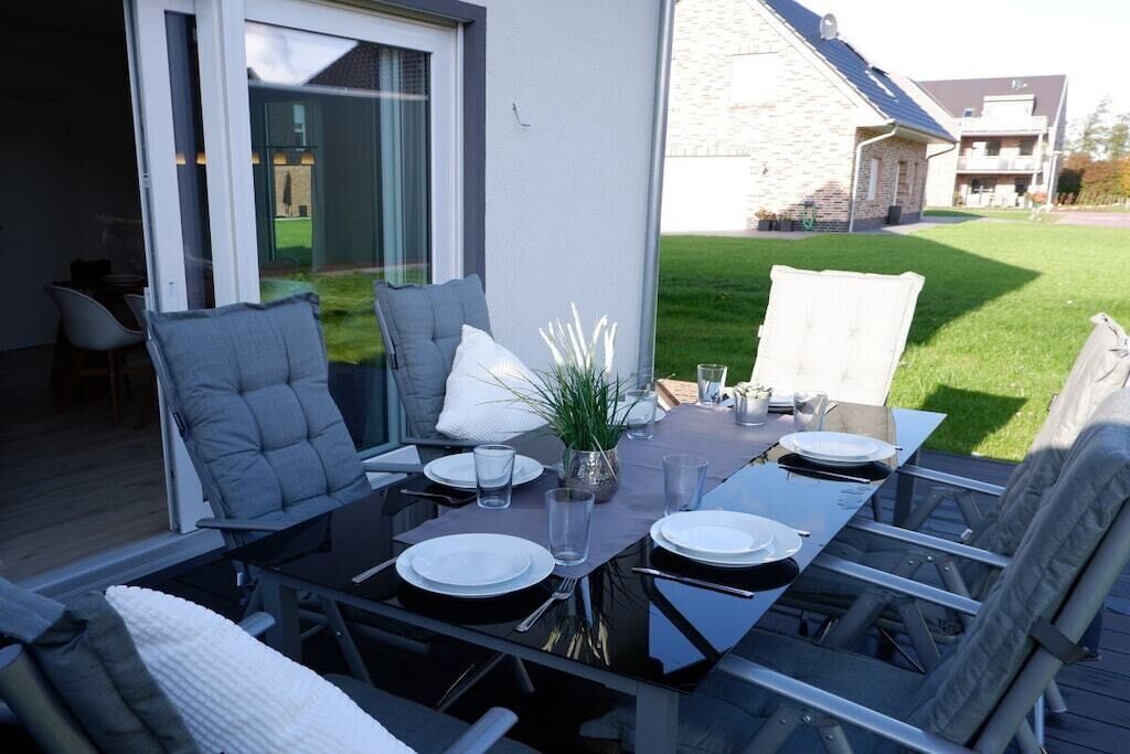 Un espace de restauration en plein air avec une table dressée pour quatre personnes, entourée de chaises, d’une plante en pot et offrant une vue sur un bâtiment et une pelouse.