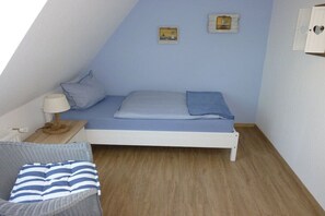 Une petite chambre avec un lit simple, un plafond en pente, une table de chevet avec une lampe, une chaise en osier et deux cadres photos accrochés au mur.