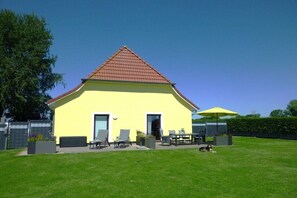 Un bâtiment jaune avec un toit rouge, une terrasse avec des meubles d’extérieur et un chien.