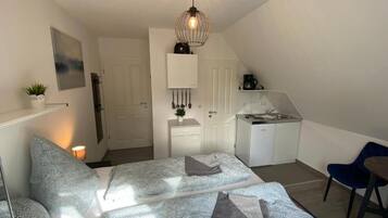 A bedroom with a bed, a desk, a chair, a hanging light fixture, and a small kitchenette.