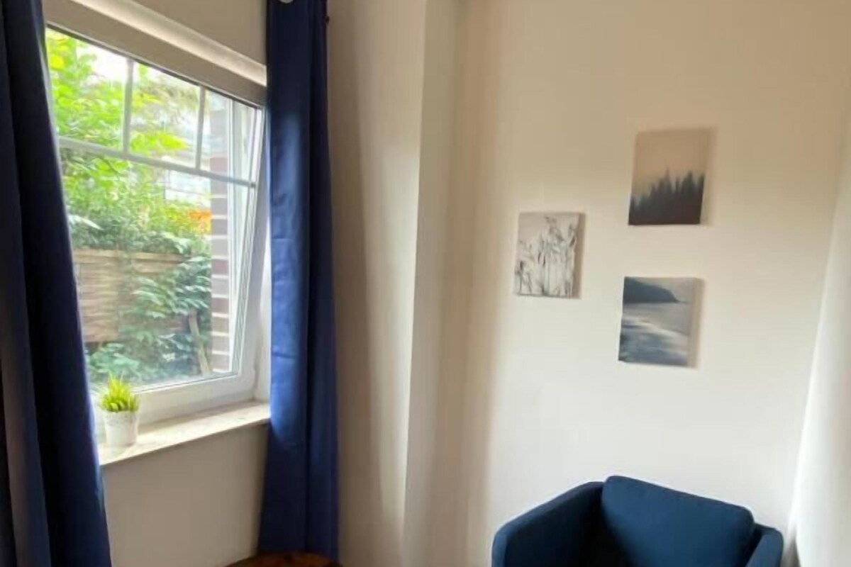 Ein Zimmer mit blauem Sessel, einem kleinen runden Tisch, einem Fenster mit Vorhängen und zwei an der Wand hängenden Bildern.